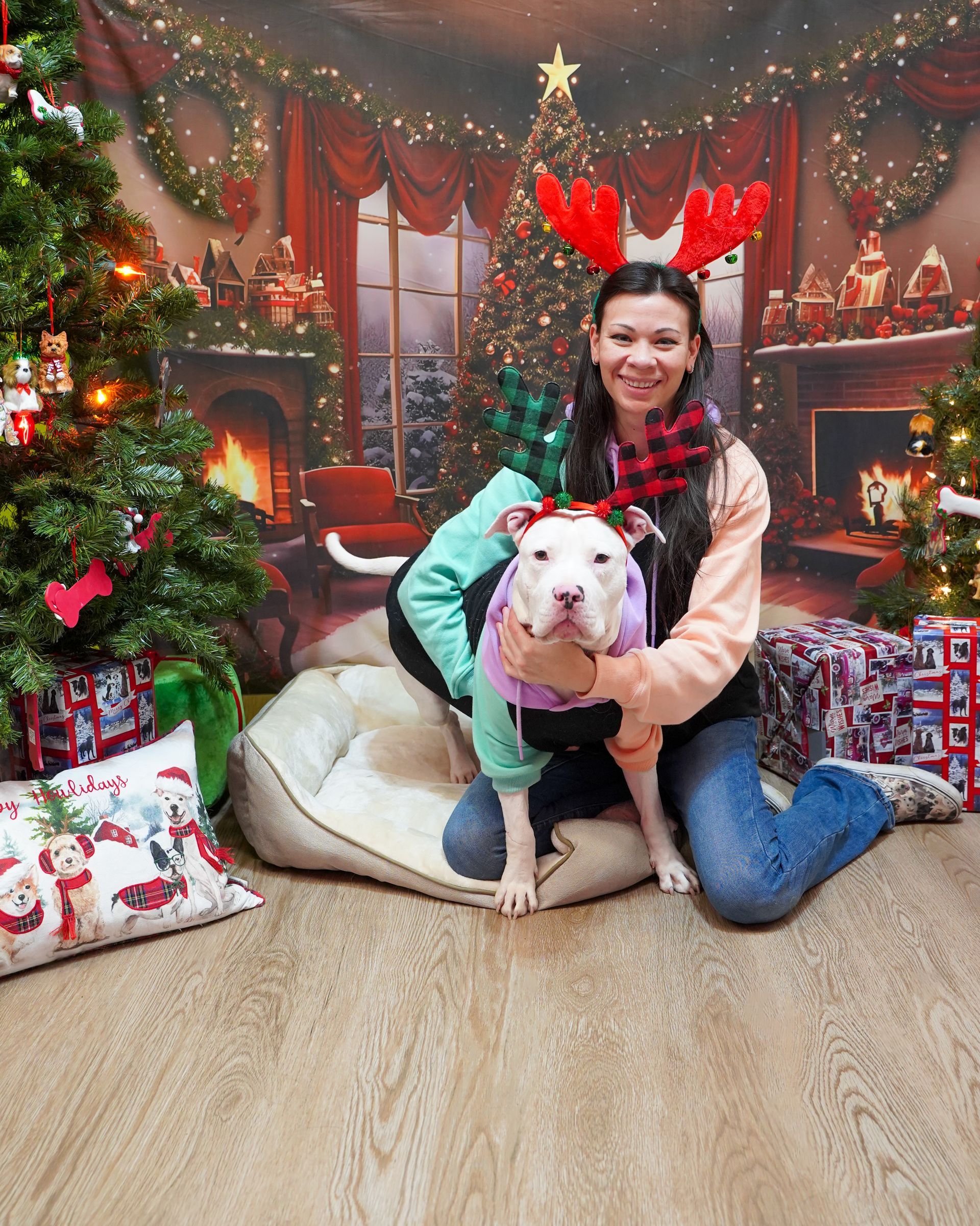 Woman and dog pose in front of a Christmas backdrop. Dog in holiday gear, woman in jeans and a scarf.