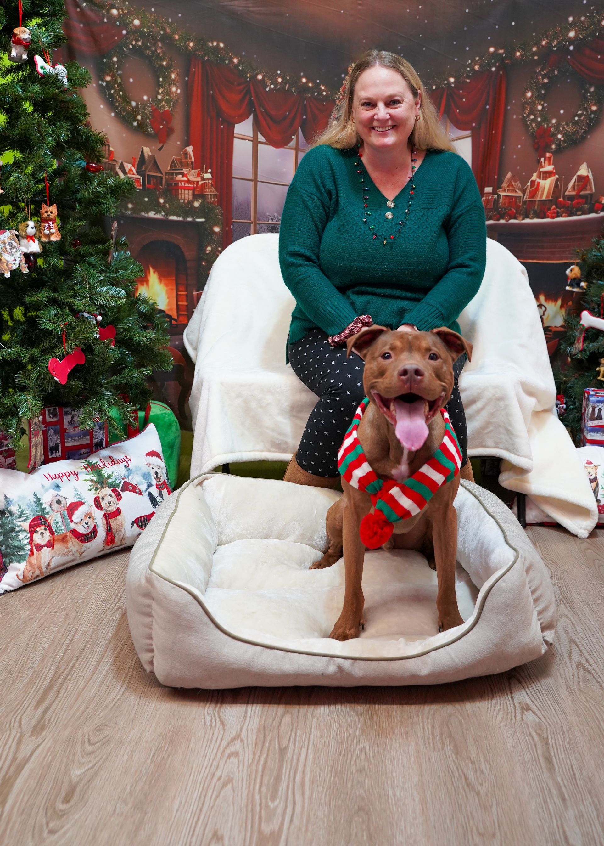 Woman and dog in Christmas setting; dog in holiday sweater sits in bed, woman smiles behind.