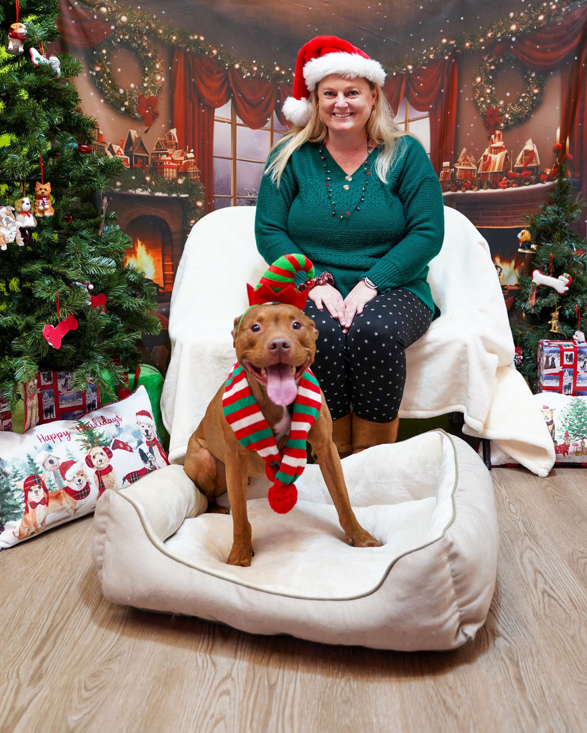 Woman in Santa hat sits with dog wearing elf hat and scarf in a holiday scene.