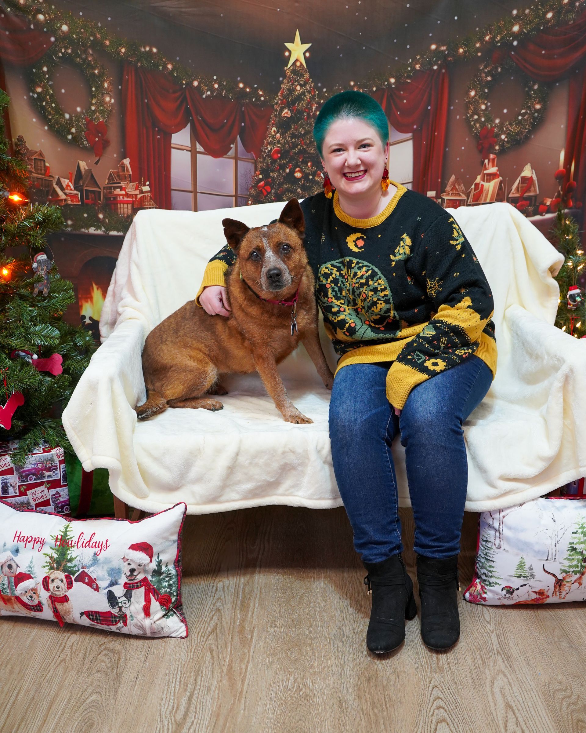 Woman with turquoise hair in a sweater, sits with a red dog on a couch in front of a Christmas backdrop.