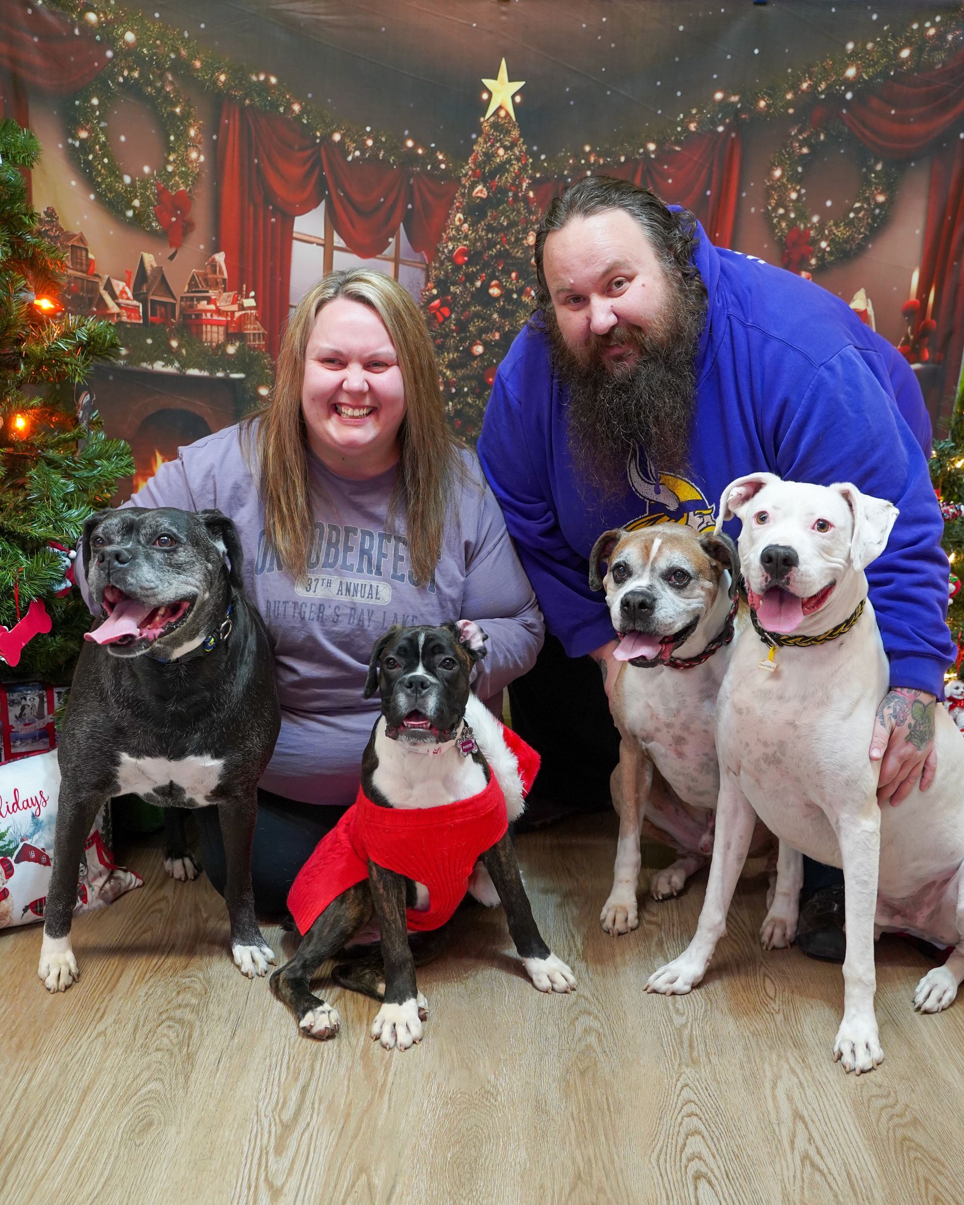 Two people pose with four dogs in front of a Christmas backdrop.