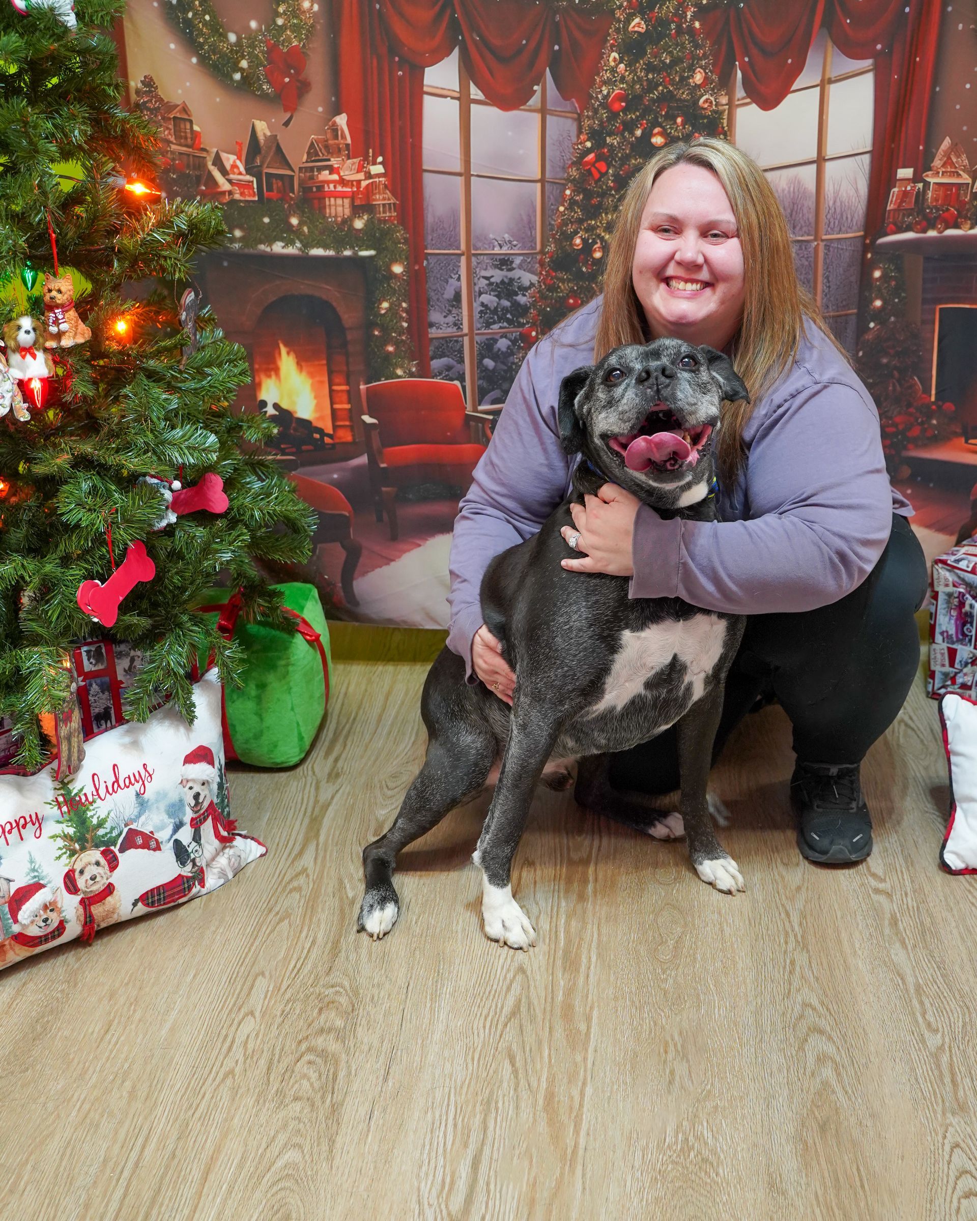 Woman with a gray and white dog posing in front of a Christmas backdrop. Woman is hugging the dog, smiling.