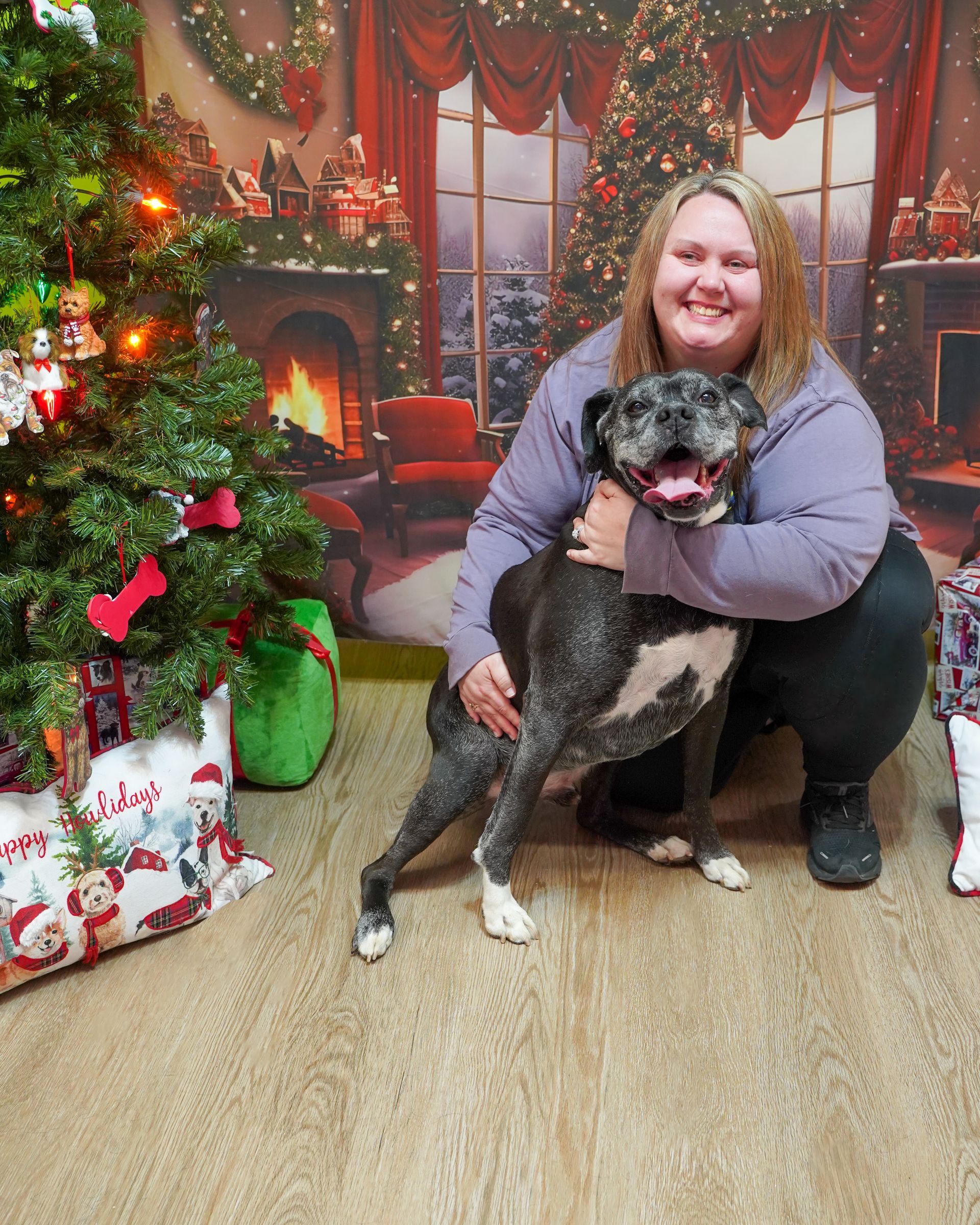 Woman hugs a spotted dog in front of a Christmas tree and fireplace backdrop.