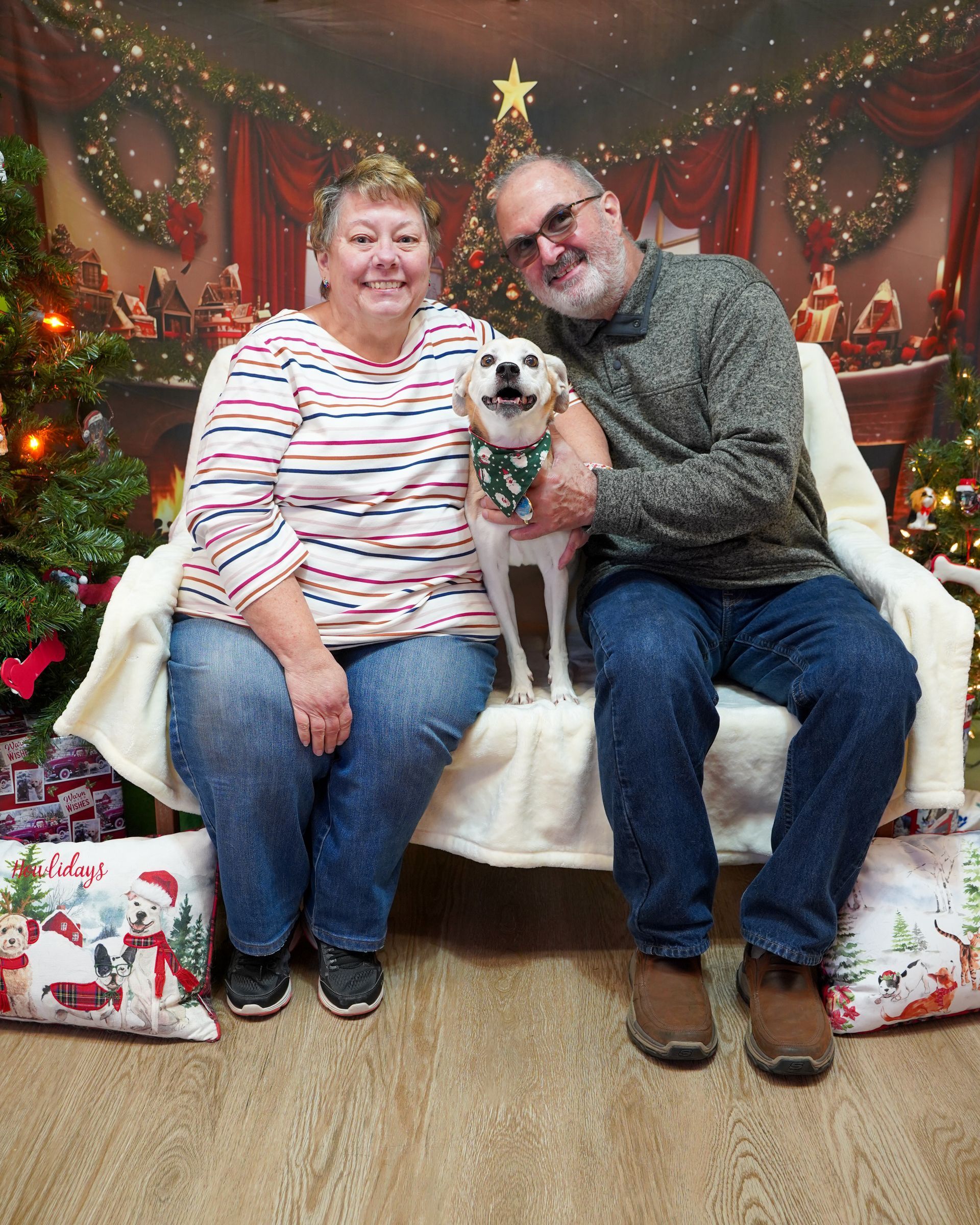 A couple sits on a couch with a dog in front of a Christmas backdrop. All smile.