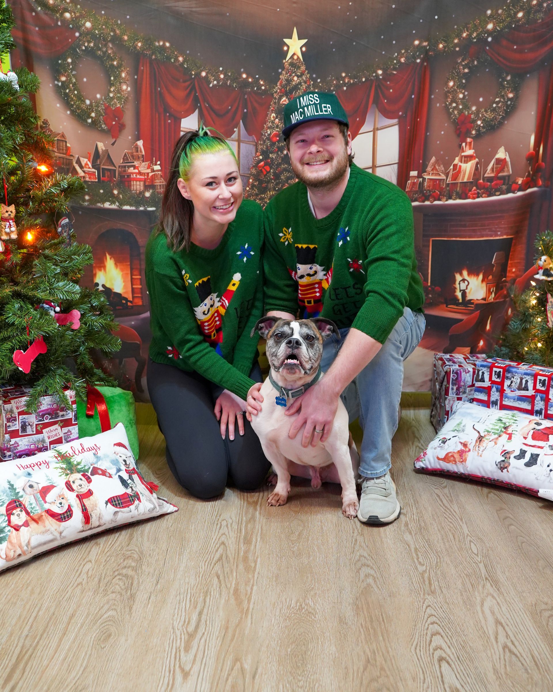 Couple and dog pose in matching Christmas sweaters, in front of a festive backdrop with a tree, fireplace, and presents.