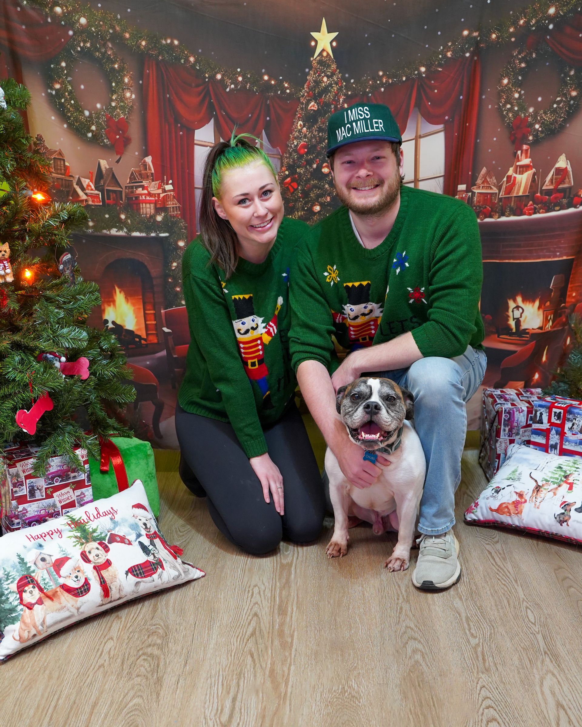 Couple and dog in matching green sweaters pose for Christmas photo. Festive backdrop, tree, presents.