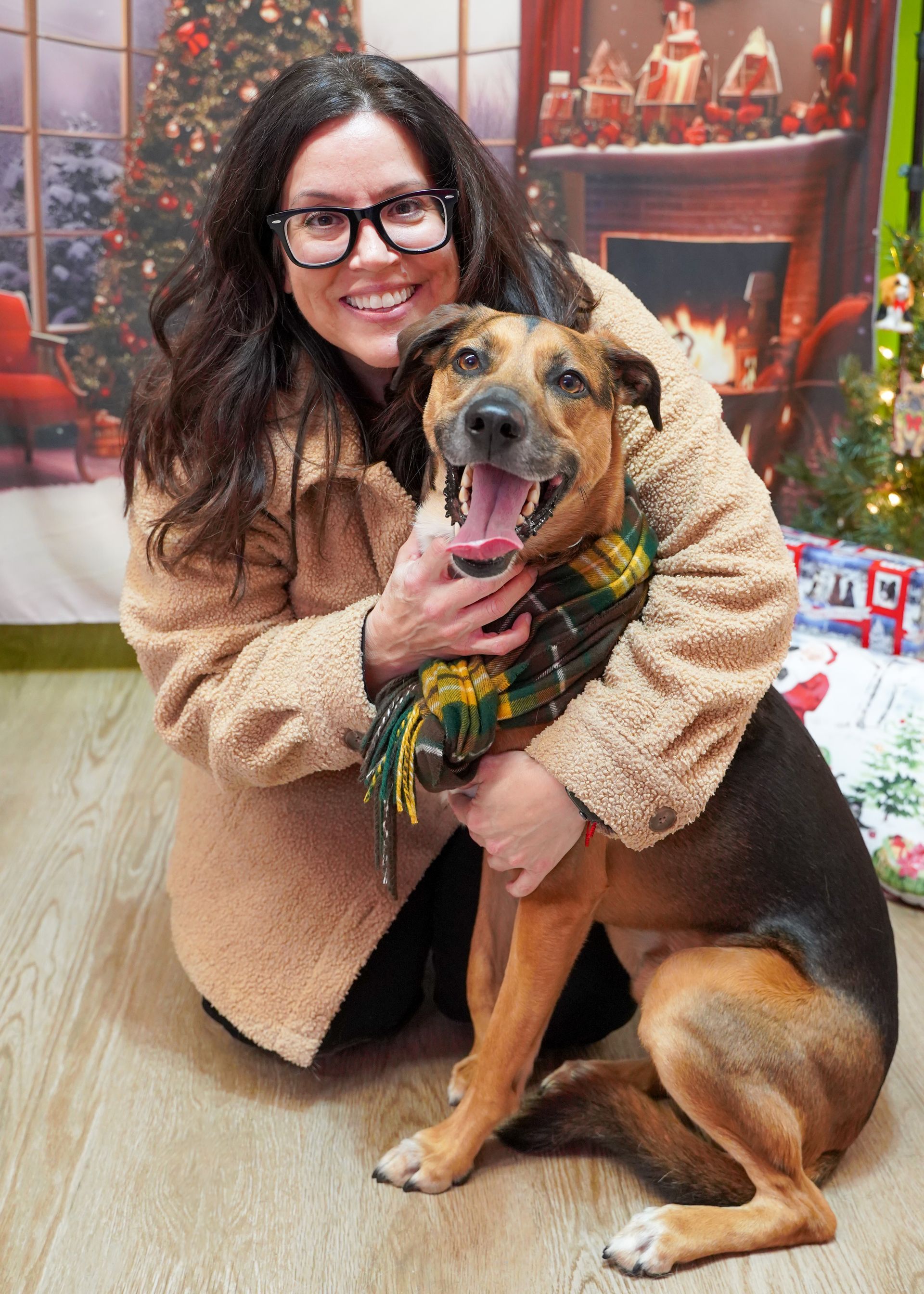 Woman in glasses hugs a dog wearing a scarf; Christmas backdrop.