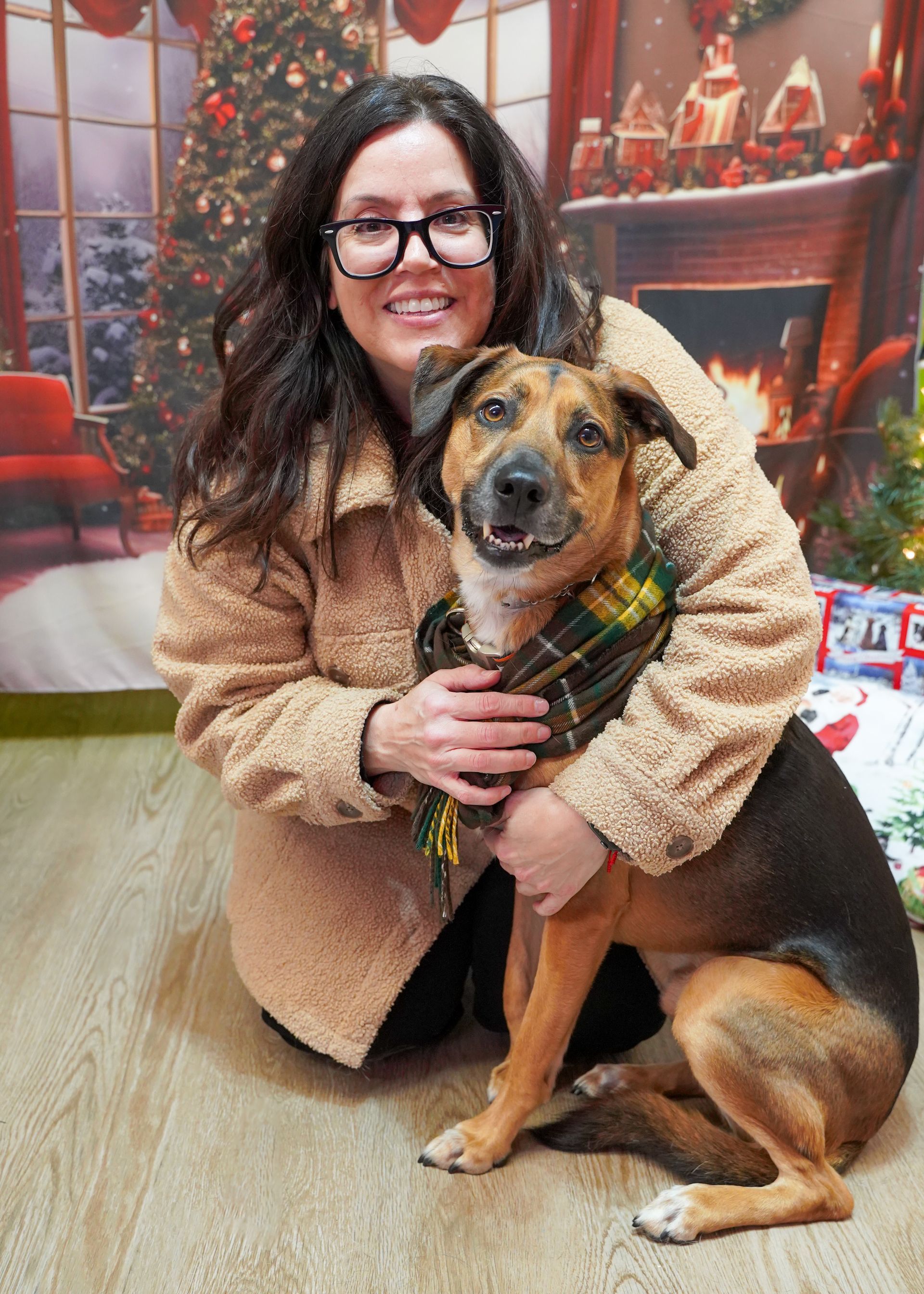 Woman with glasses hugging a dog wearing a scarf in front of a Christmas backdrop.