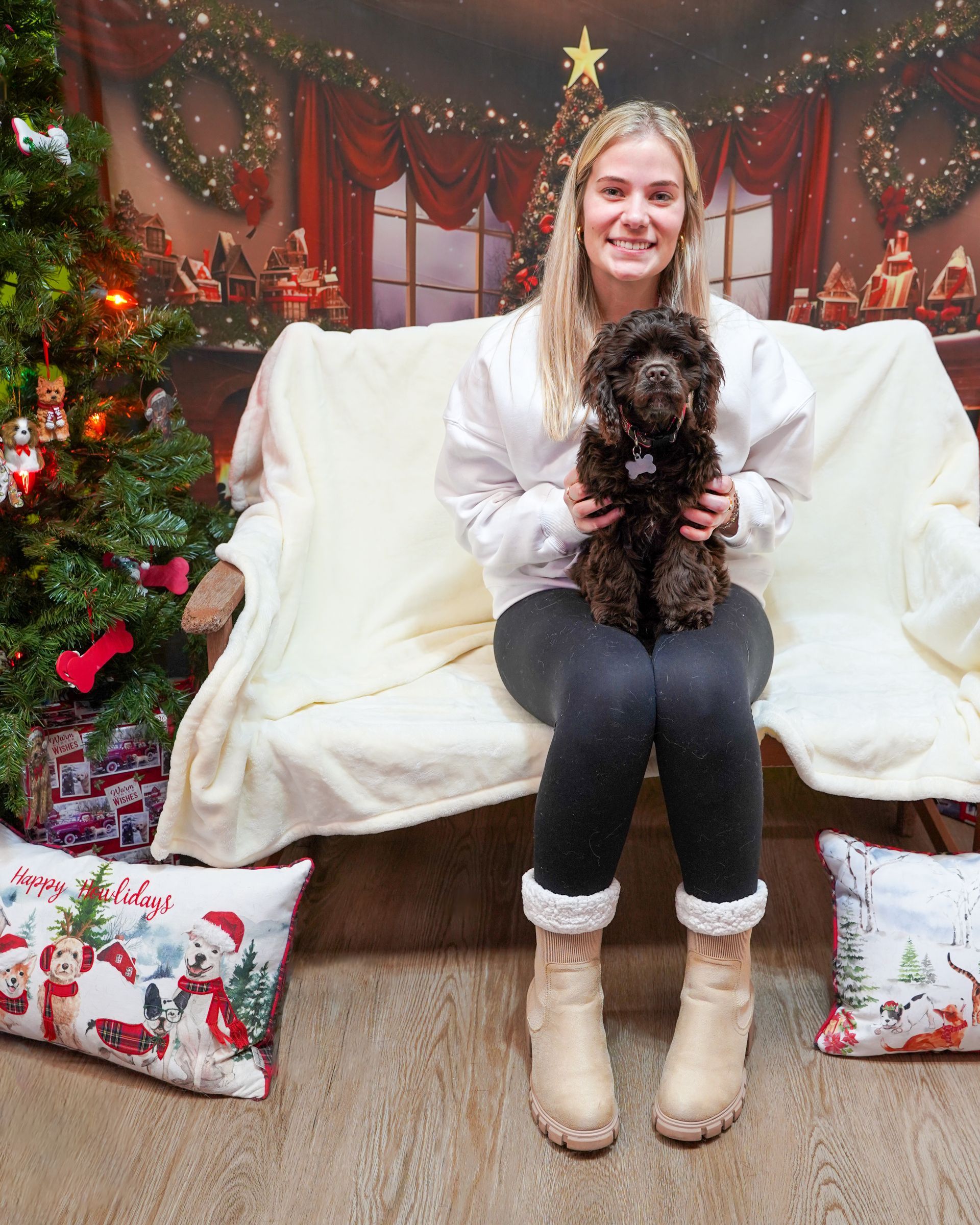 Woman holding a small black dog on a couch in front of a Christmas backdrop.