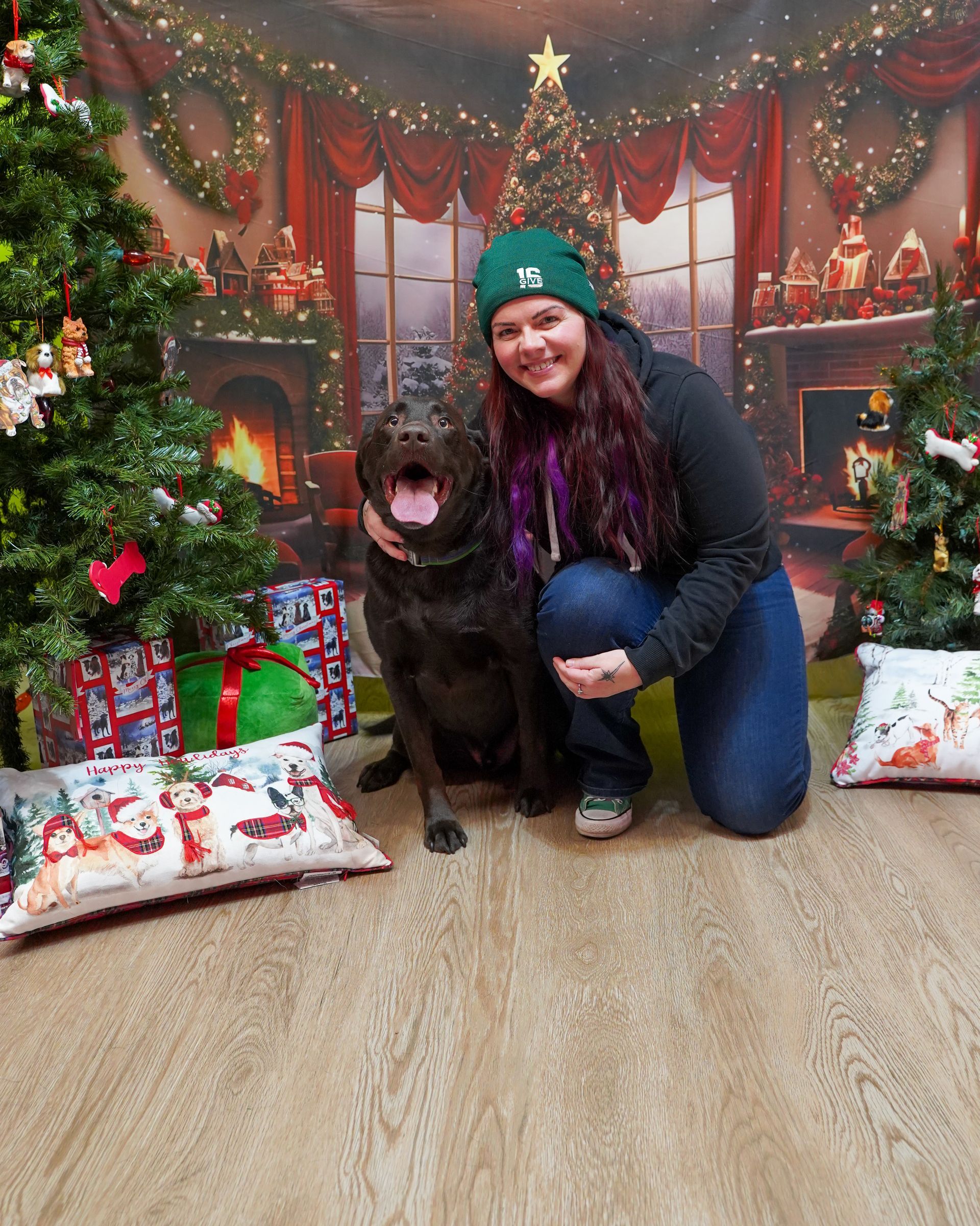 Woman kneels beside a chocolate lab in front of a Christmas backdrop. They smile near a decorated tree.