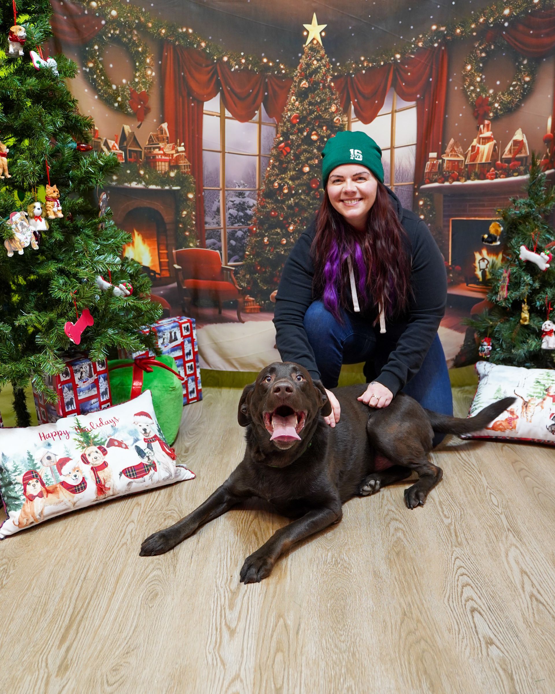 Woman and chocolate lab pose for Christmas photo in front of a festive backdrop.