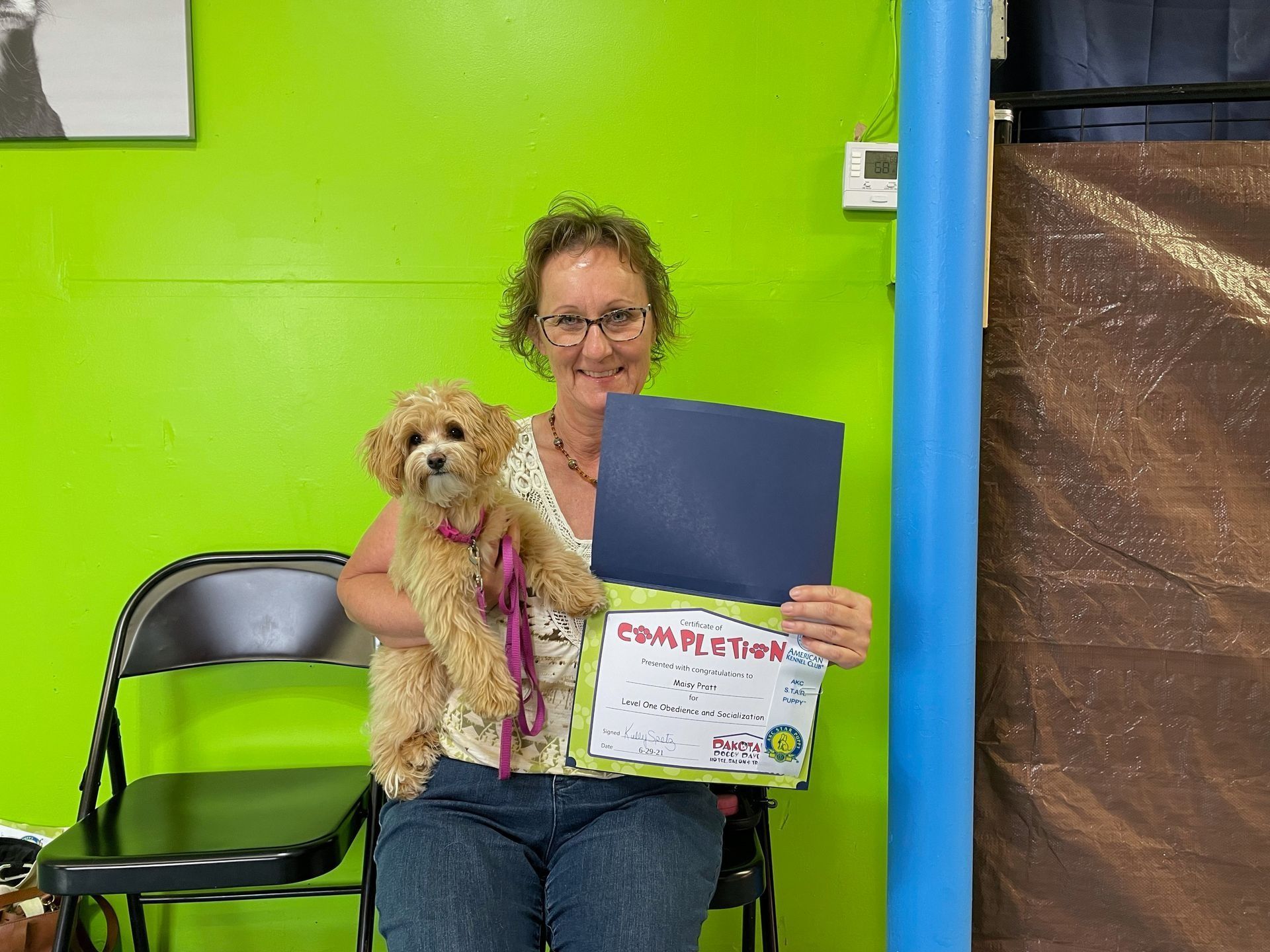 Woman seated with a small dog, holding a certificate. They are in front of a green wall.