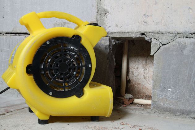 Yellow portable blower fan beside a cinder block wall near a damaged opening.