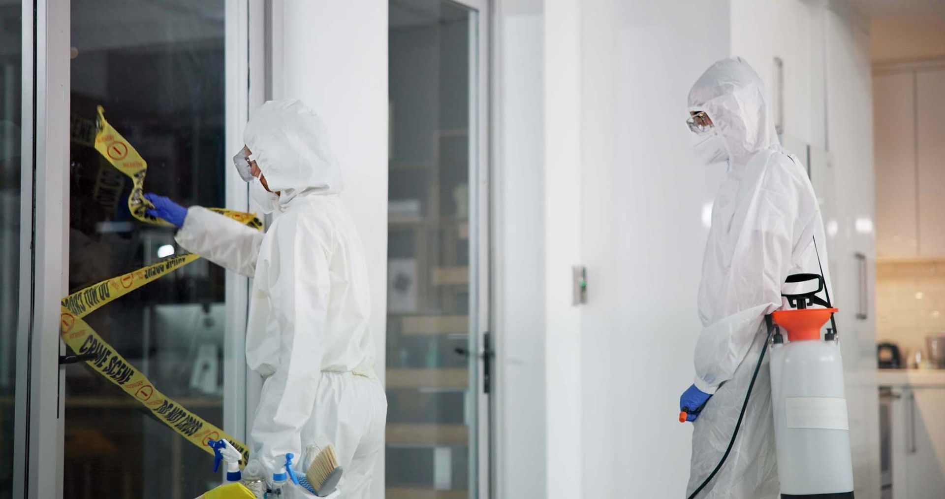 Two workers in white protective suits inside a building, one wrapping yellow caution tape around a doorway.