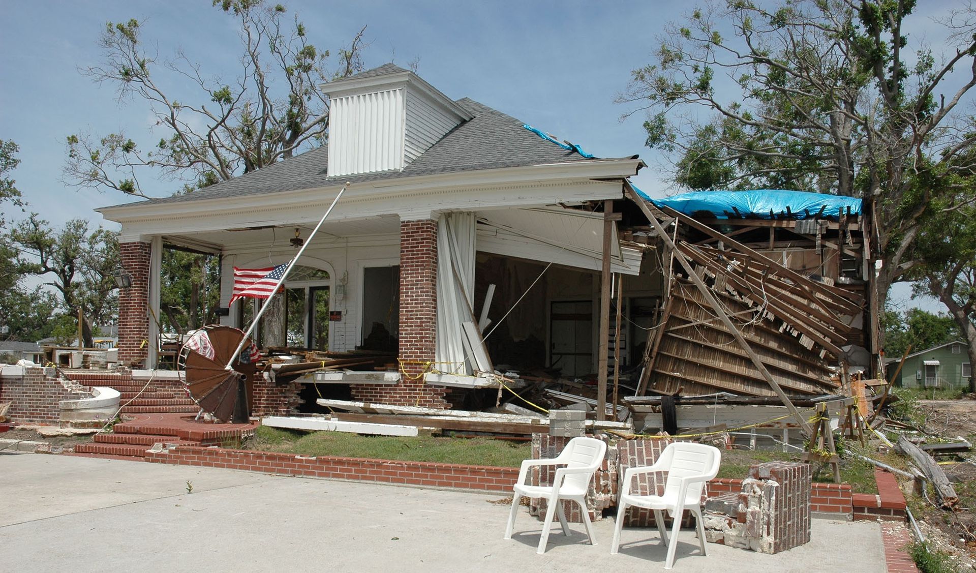 Damaged house with collapsed roof and debris after a storm, with chairs and an American flag out front