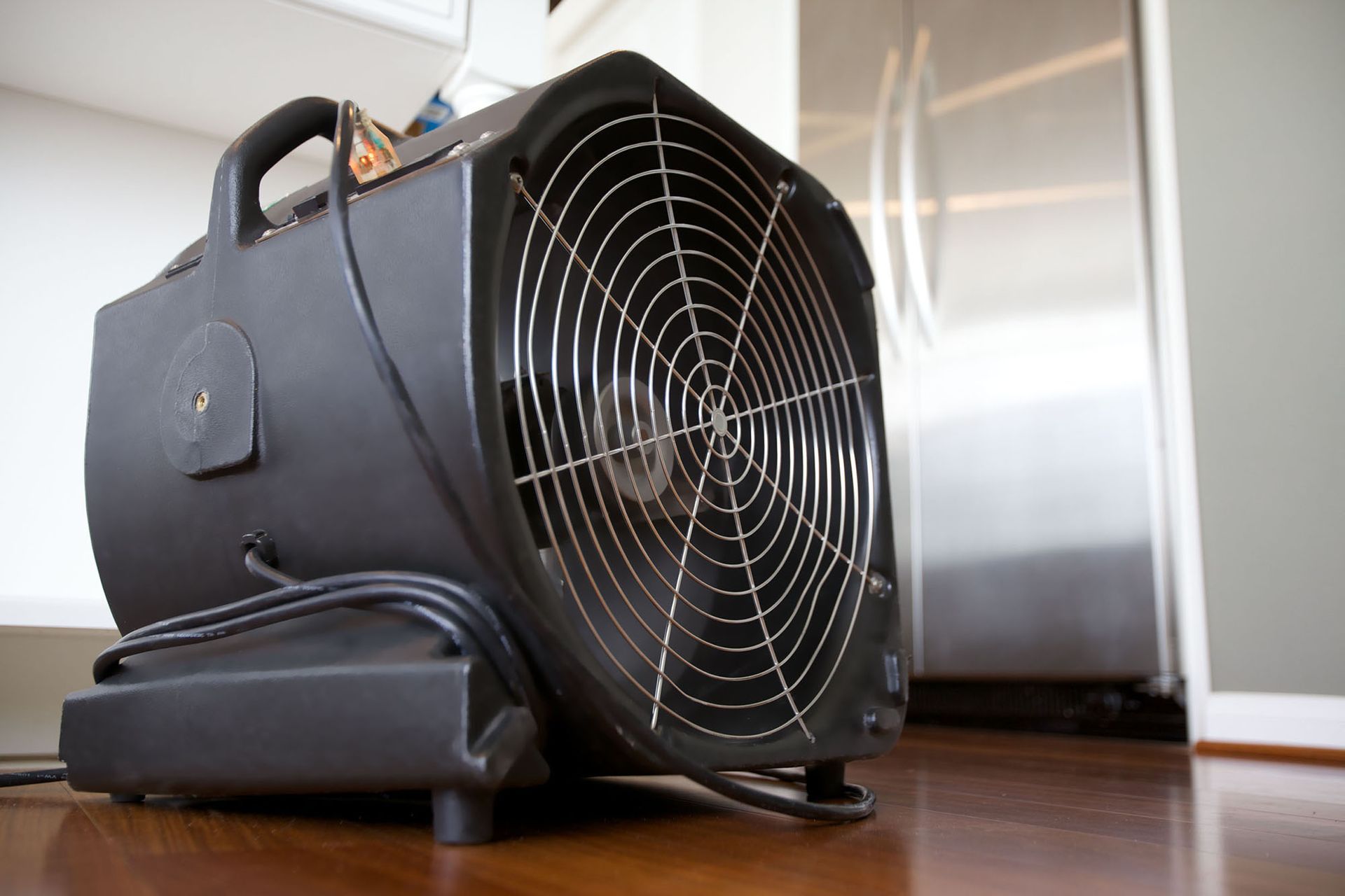 Portable black floor fan on a wooden floor in a kitchen or hallway, viewed from the side.