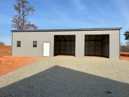 Metal storage building with two open bays, a white door, and gravel driveway under clear sky.
