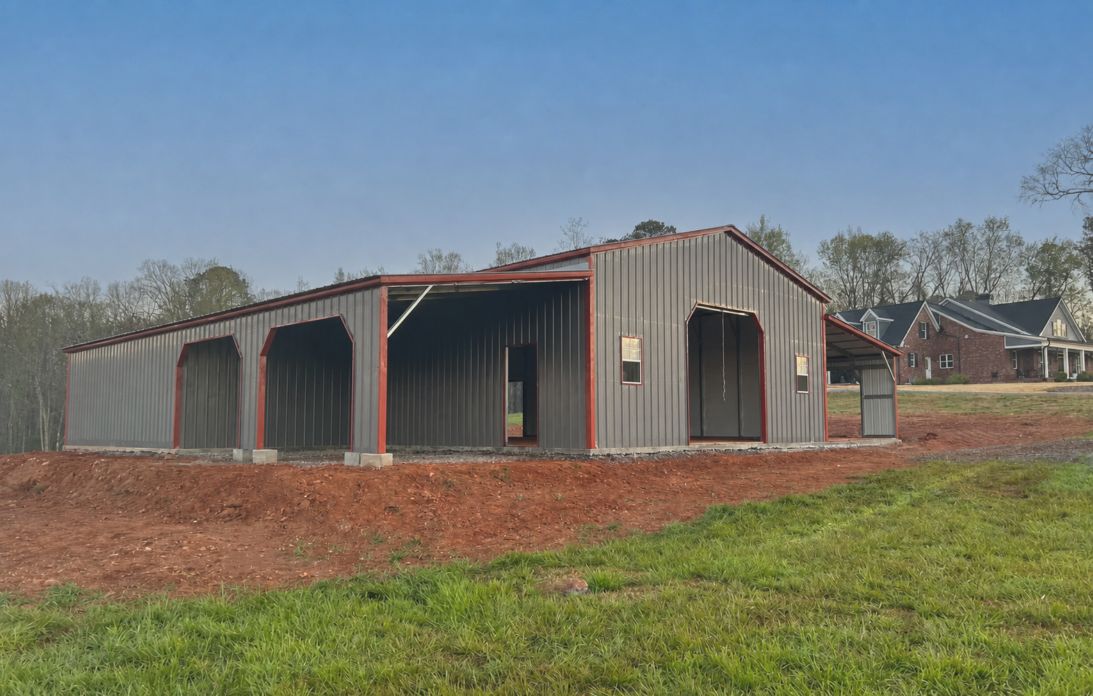 Large gray metal barn with red trim on a grassy rural lot under a clear sky