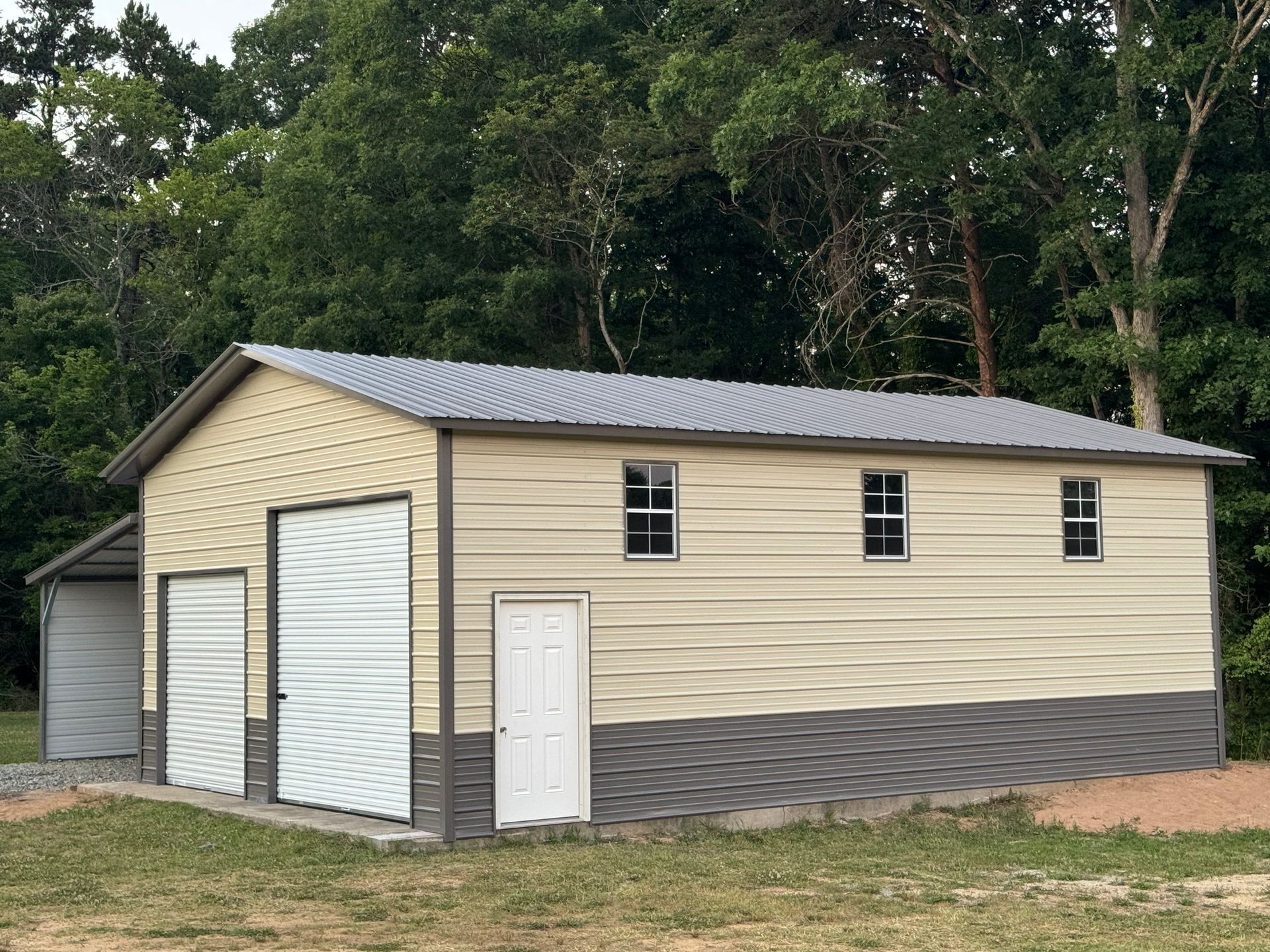 Tan metal garage with beige siding and three doors, set beside trees on a dirt lot