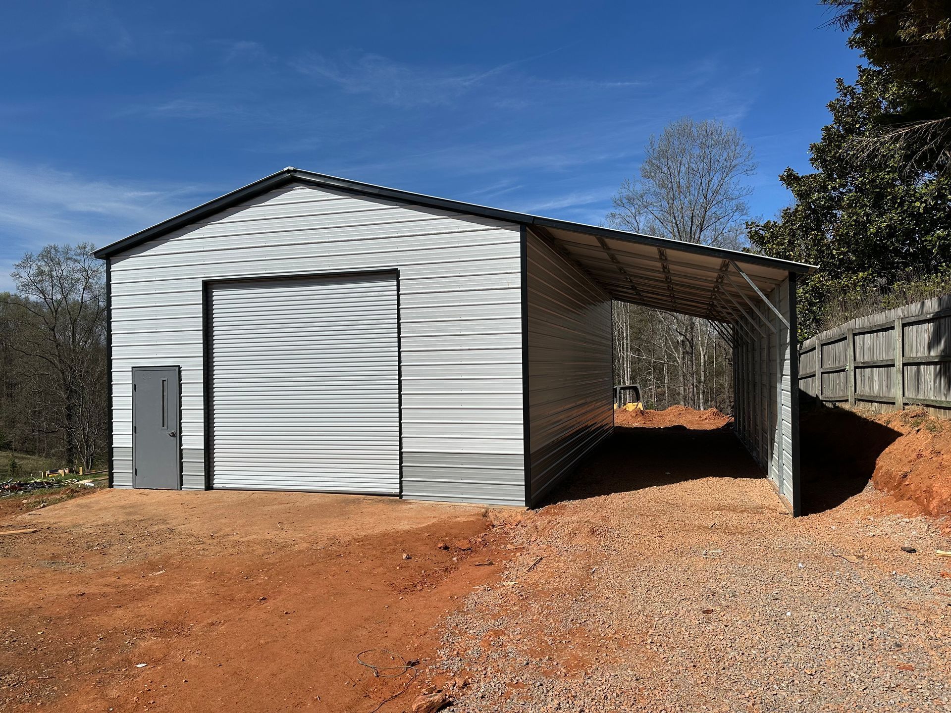 White metal garage with attached open carport on a red dirt lot under a blue sky