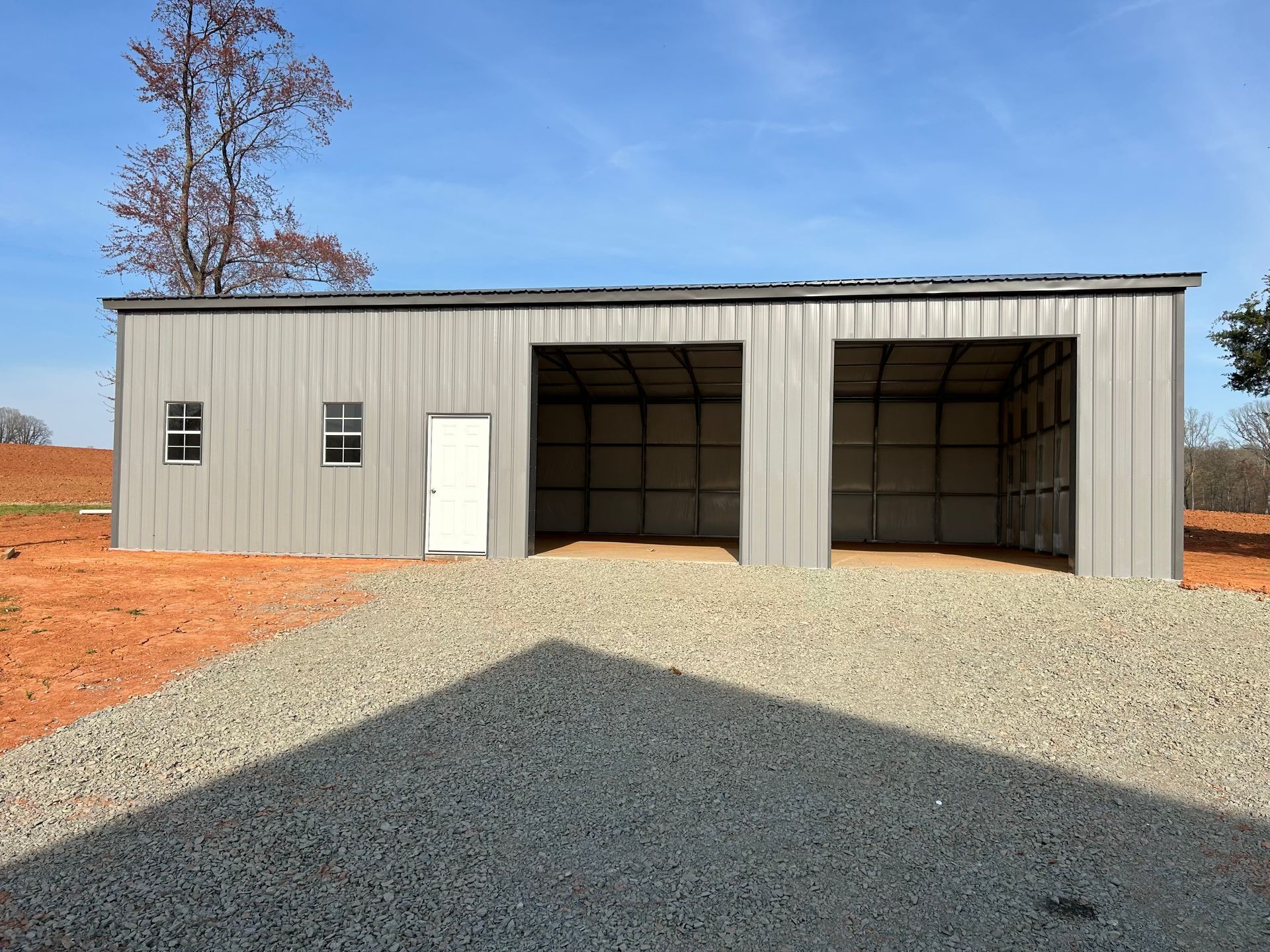 Metal building with two open garage bays and a white door, set on gravel under a clear sky.
