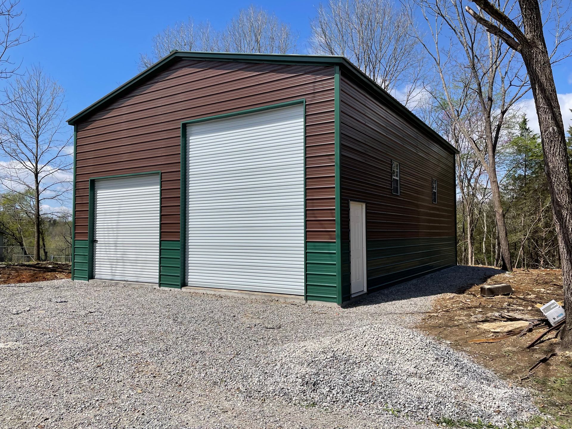 Brown metal garage with two white doors on a gravel lot, surrounded by trees and blue sky