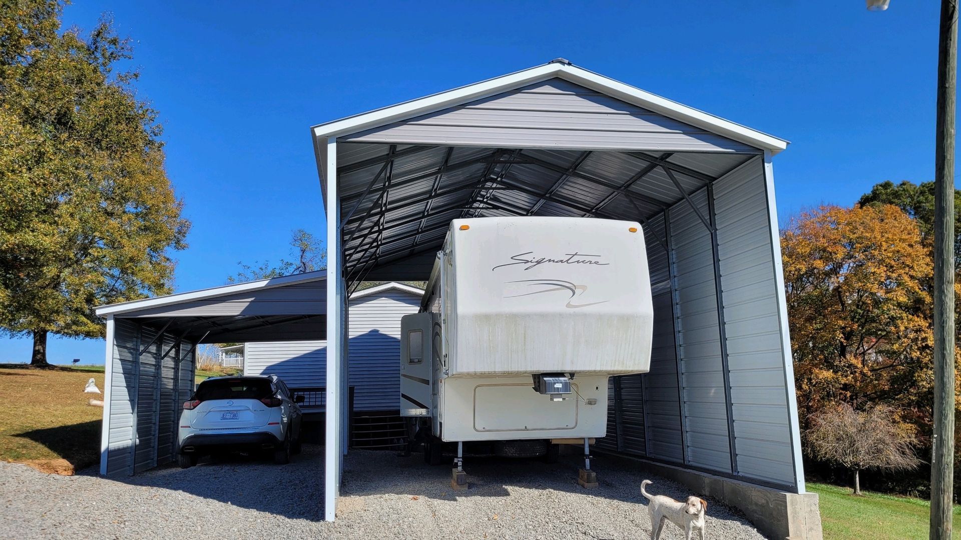 White covered parking shelter with a car and large white RV/trailer parked inside on a sunny day