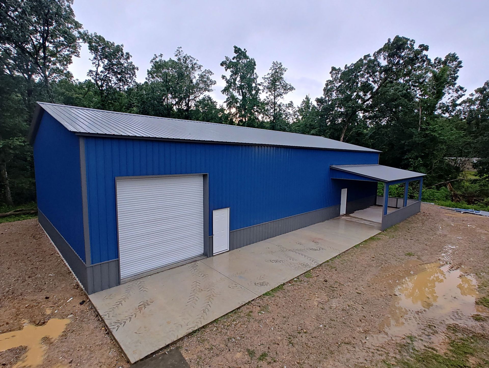 Blue metal building with white doors and awning beside a concrete pad on a gravel lot