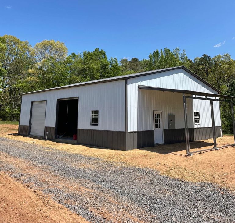 White metal barn with open bays and covered porch on a gravel lot, surrounded by trees under a blue sky
