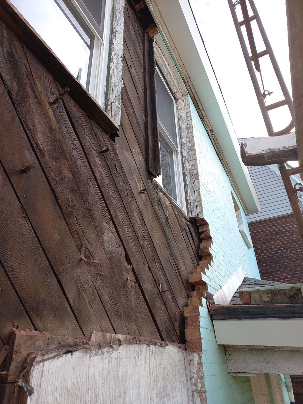 Exterior view of a building with wooden siding, showing areas of damage and deterioration near a window. Brown and white colors are prominent.