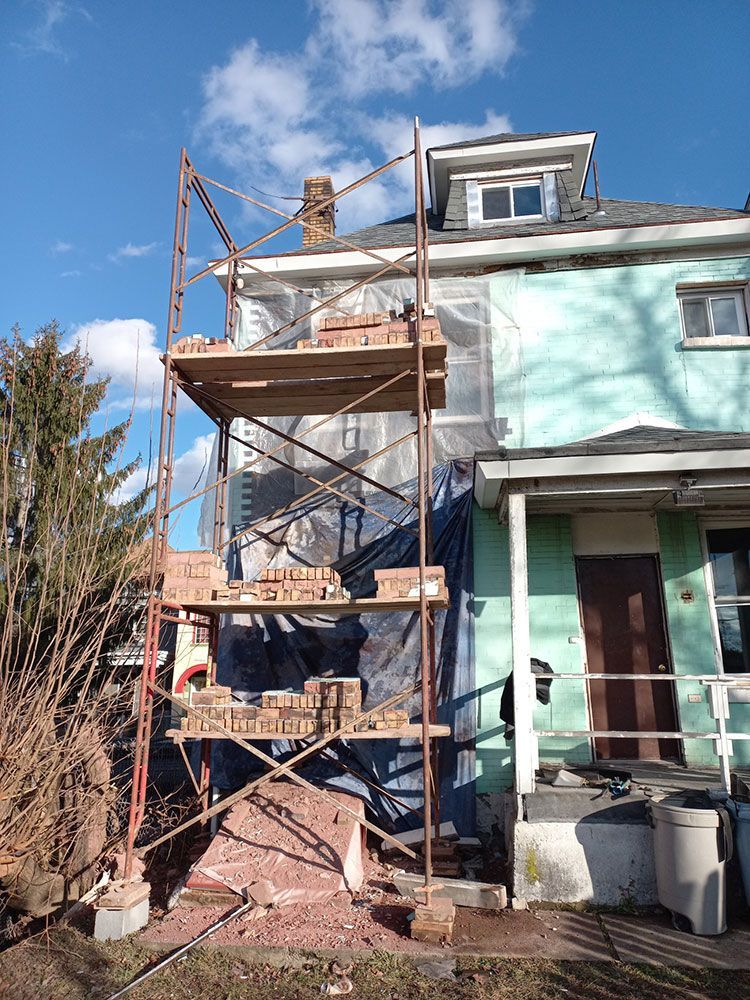 Scaffolding set up against a house with brickwork visible. The house exterior is a mix of blue and green, with a chimney at the top.