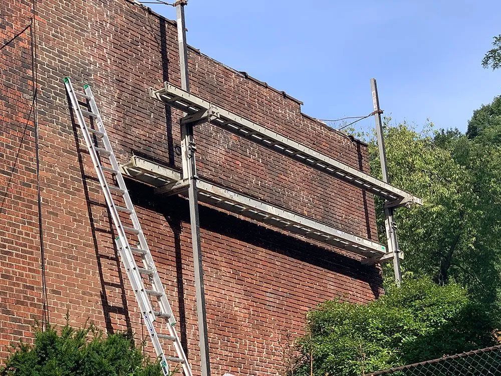 A tall brick wall with scaffolding and a ladder set up. Green bushes are along the bottom and trees in the background.