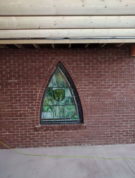 Brick wall with a Gothic-shaped stained-glass window featuring a green shield design. Wooden beams and a concrete floor are visible.