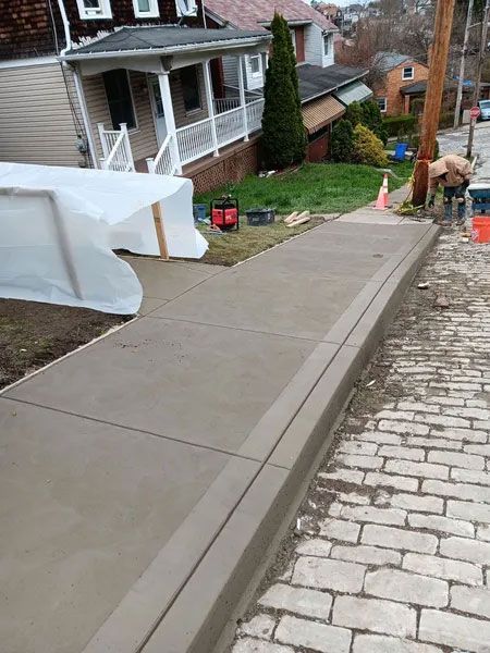 Newly poured concrete sidewalk with curb alongside a brick-paved street. A worker is visible in the background.