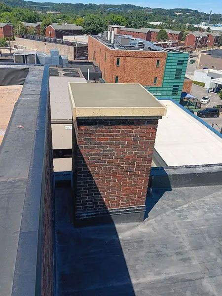 Brick chimney on a flat roof with a beige cap; surrounding rooftops and buildings in the distance under a blue sky.