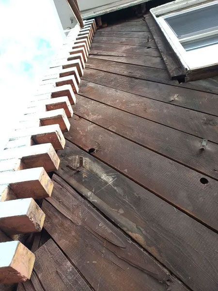 Exterior view of a weathered wooden wall with a row of brick-like structures running vertically along its edge. A window is visible.