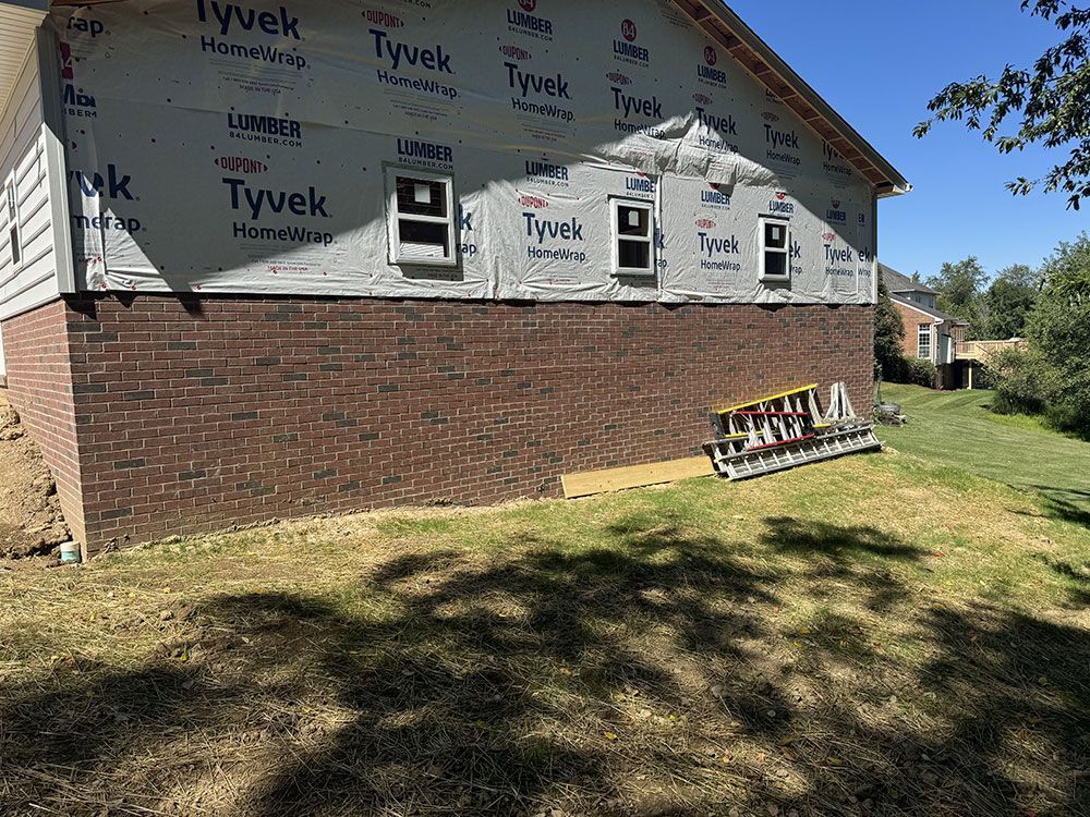 Brick home exterior under construction with Tyvek wrap and three windows above. Green grass in foreground.