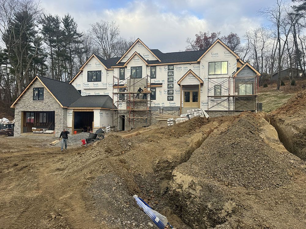 House under construction with workers, surrounded by dirt and trenches. Exterior walls are wrapped in paper, with stone and wood accents.
