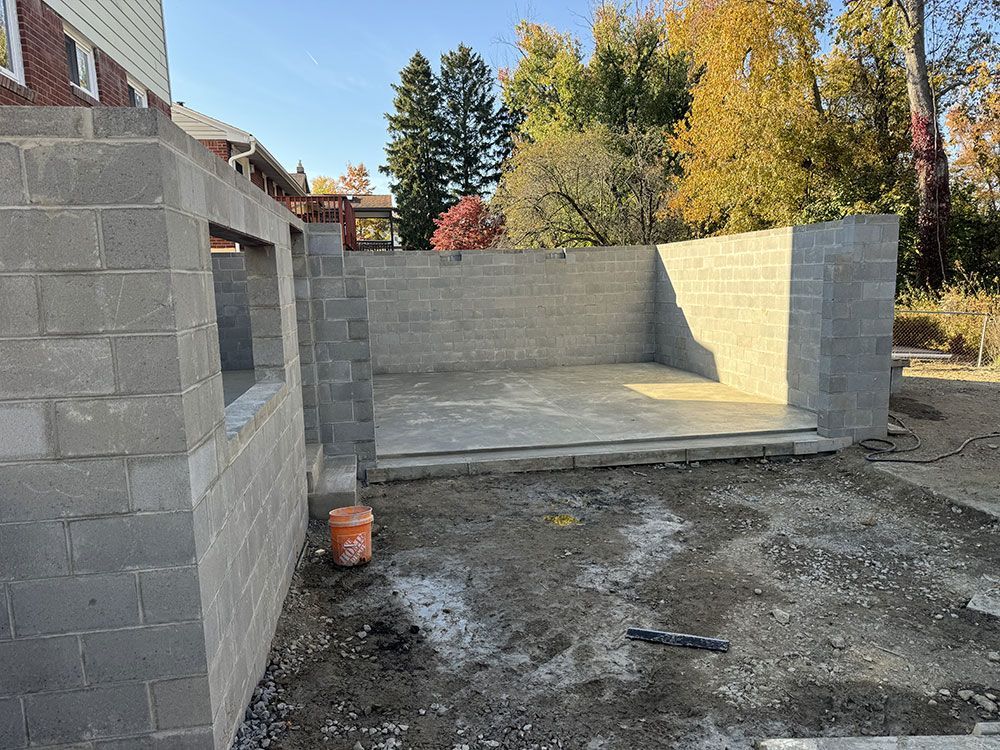Construction site: concrete block walls and concrete floor under construction, with a partially constructed building surrounded by dirt, with trees in the background.