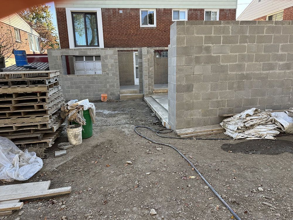 Construction site with gray cinder block walls, a concrete slab, and debris scattered on the ground; a red brick building is in the background.