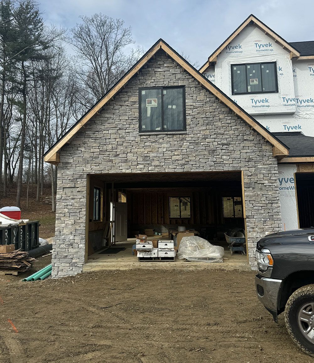 Garage under construction with gray stone facade, beige trim, and a truck parked to the right. The house is in a wooded area.