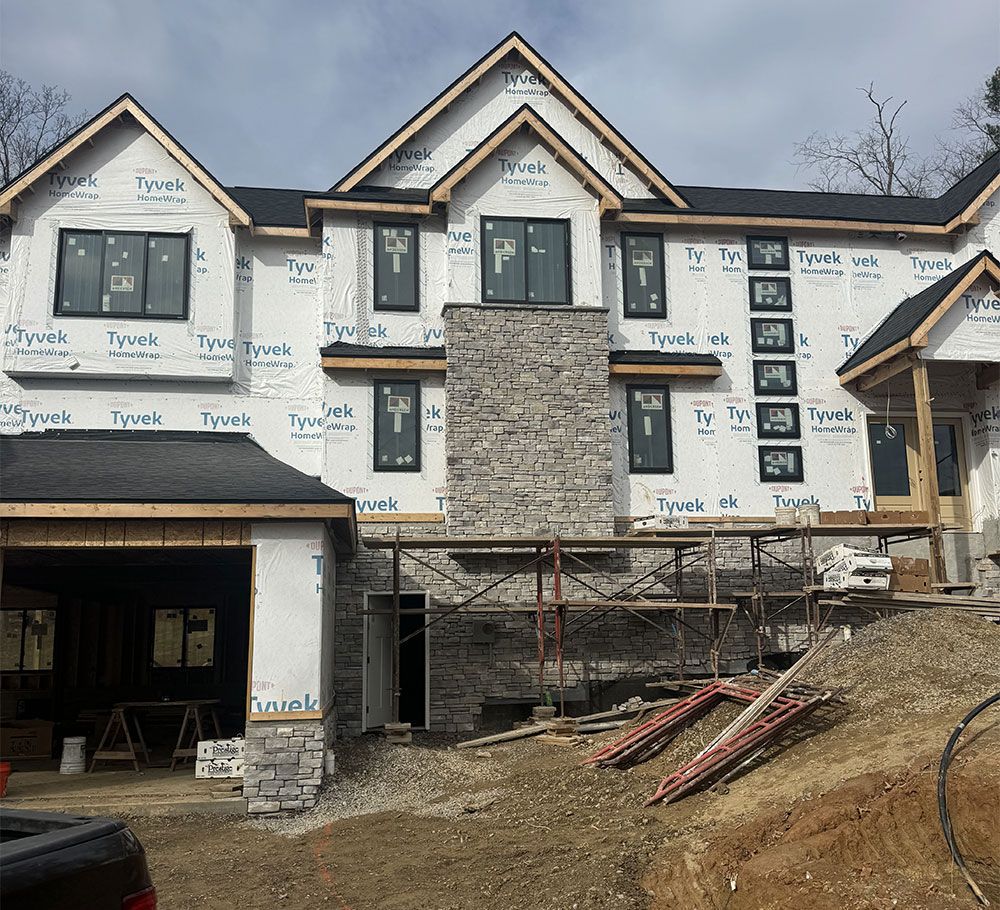 Two-story house under construction with stone facade, Tyvek wrap, and black window frames. Scaffolding and building materials are visible.