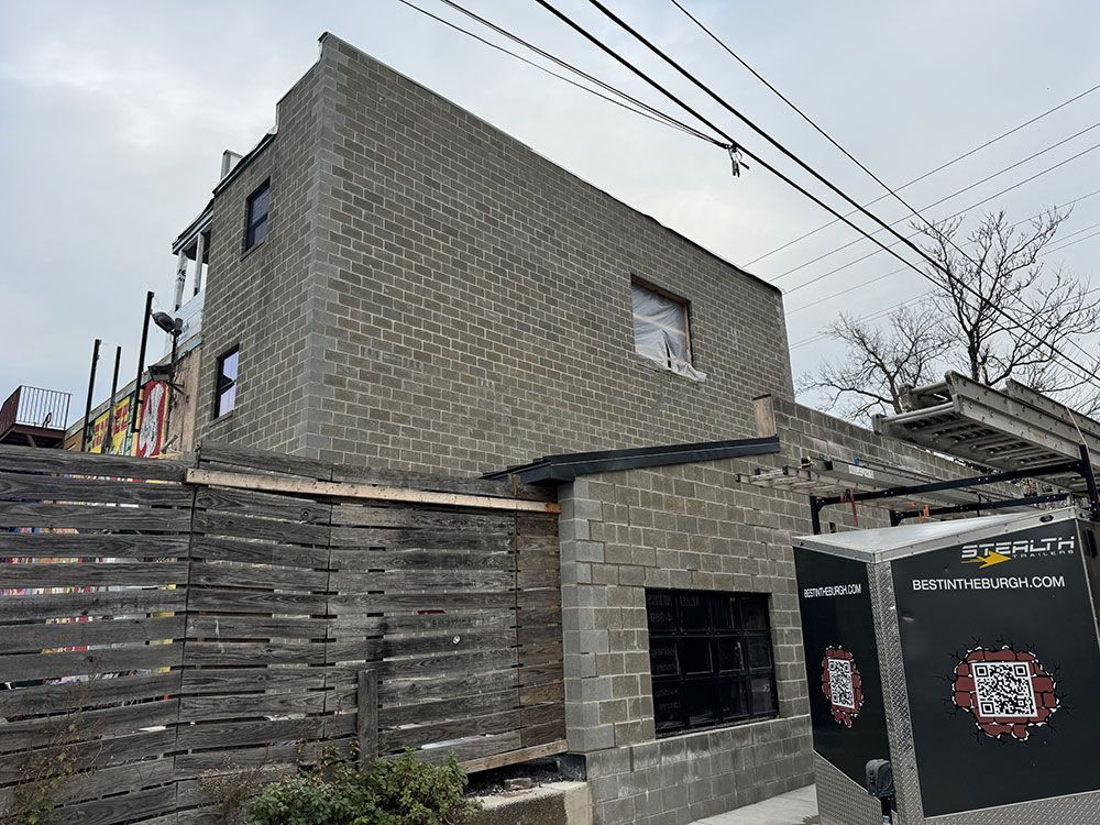 A two-story building under construction with concrete block walls. A black box with a QR code stands in front of a wooden fence.