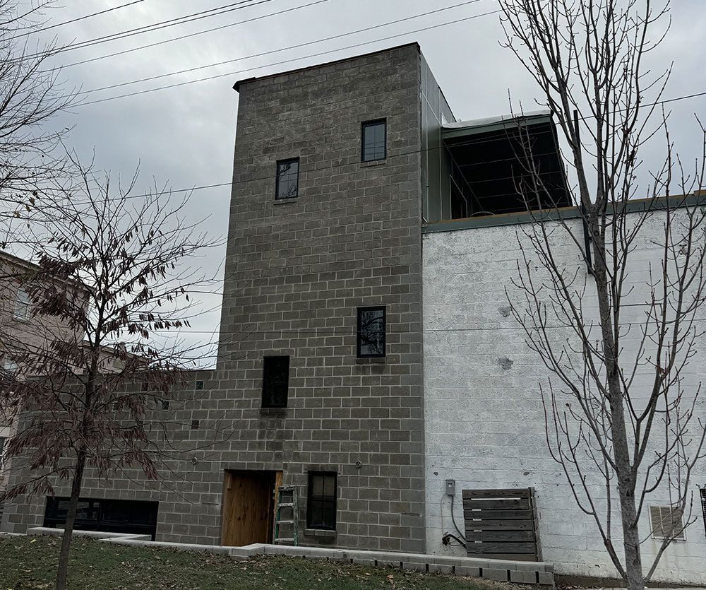Exterior view of a multi-story brick building with dark rectangular windows. The side wall is painted white. Bare trees are in the foreground.