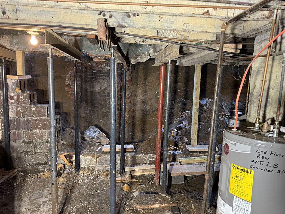 Basement view with exposed brick, support beams, and a water heater. Metal jacks support the ceiling, suggesting structural work is underway.