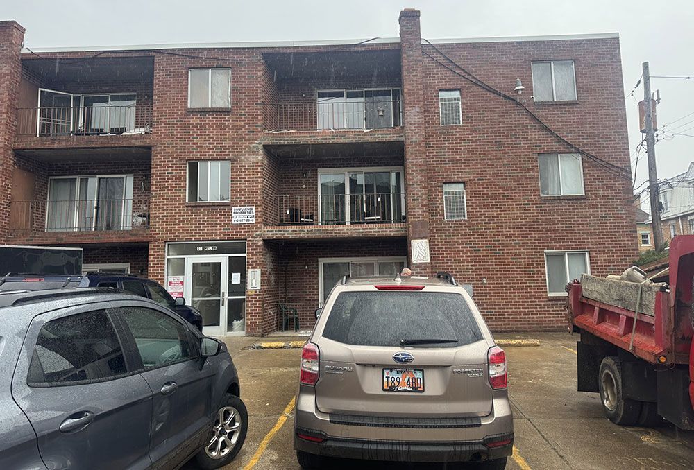 Exterior of a three-story brick apartment building with balconies. Cars are parked in front.