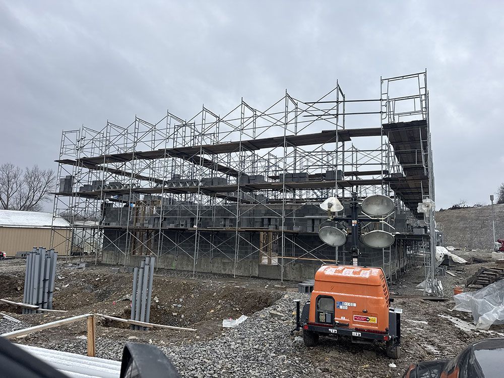 Construction site with scaffolding around a dark structure, featuring an orange generator in the foreground and a cloudy sky.