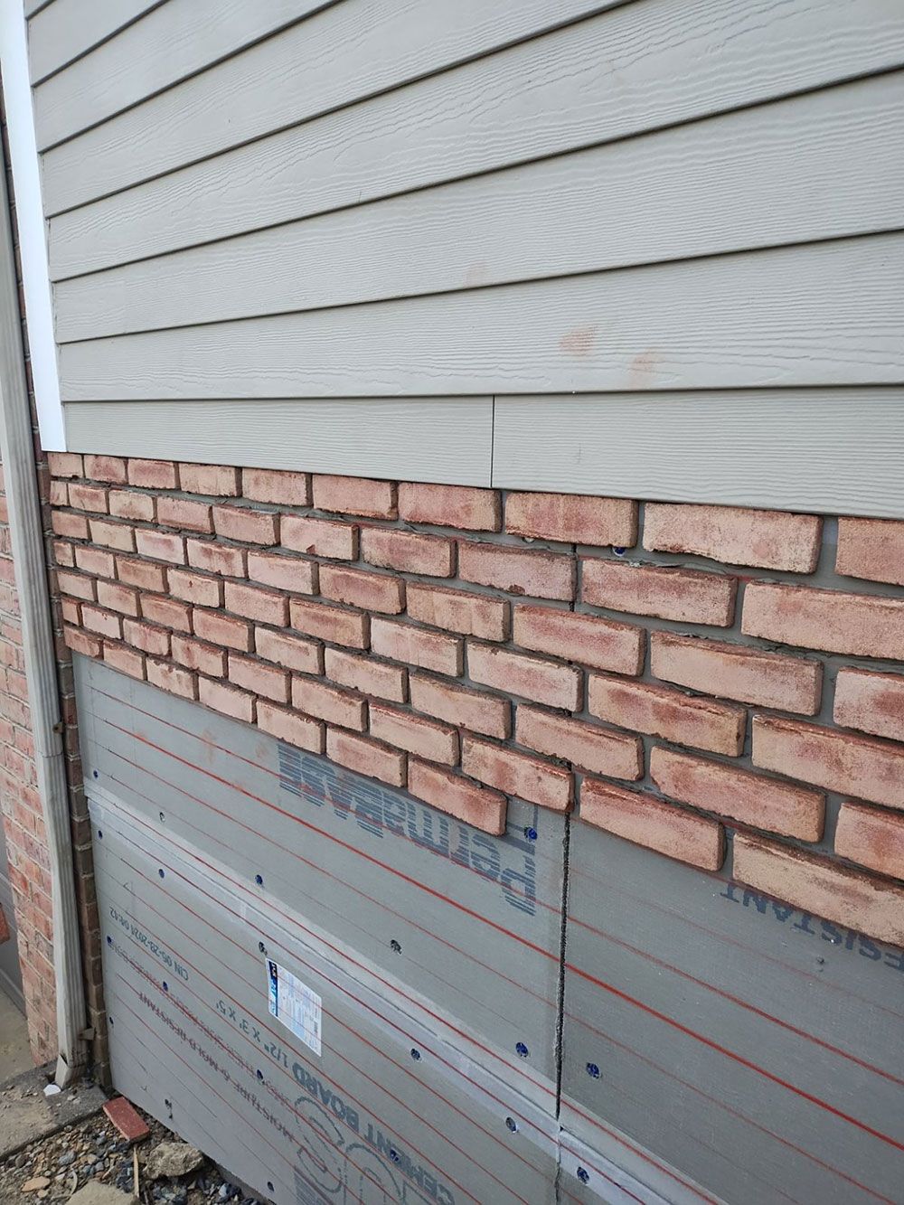 Construction of a building exterior with gray siding, brick veneer, and a gray underlayment visible.