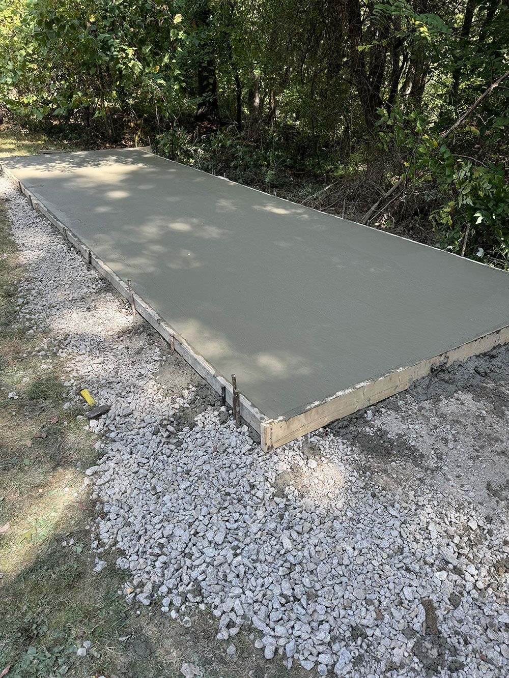 Freshly poured concrete slab, rectangular, framed by wooden forms, surrounded by gravel, in a natural outdoor setting with trees.