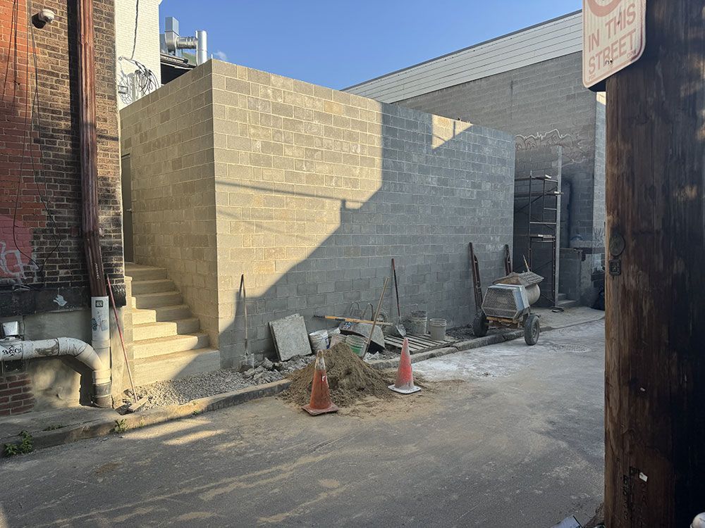 Construction in an alleyway. A gray cinder block structure with stairs dominates the scene. Debris and construction materials litter the ground.